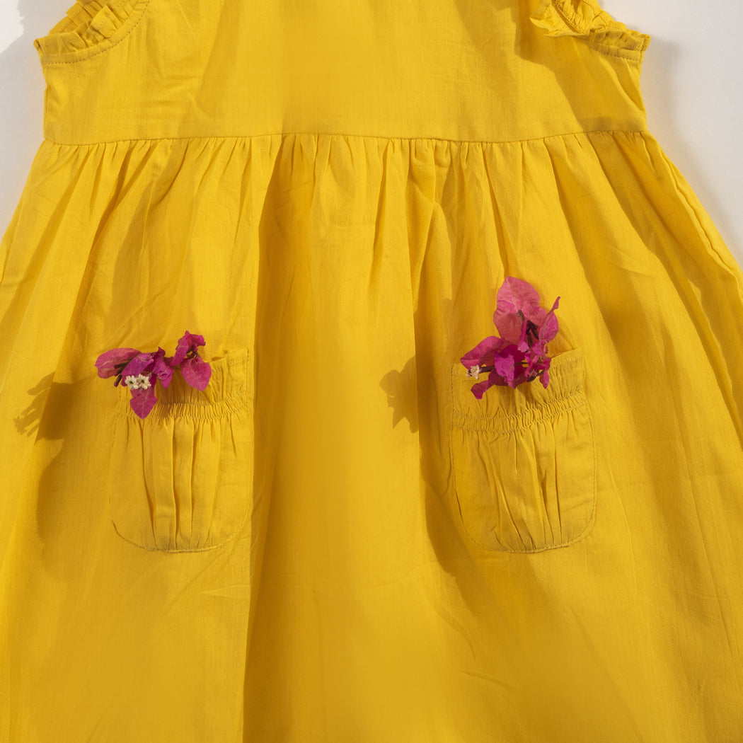 A sunshine yellow colored sleeveless dress for kids in cotton mulmul. This mid length dress has front pockets and a back button fastenings, accentuated with ruffles on the sleeve, neck and pockets.