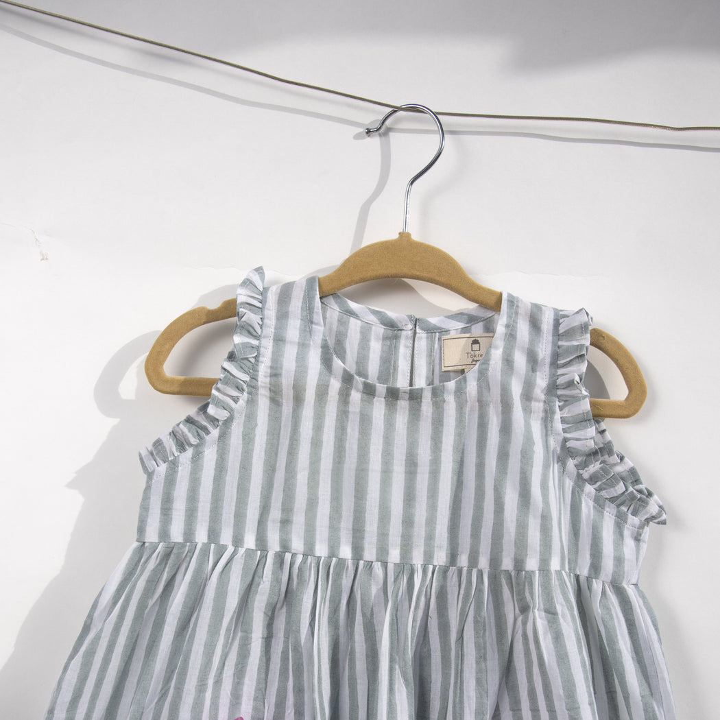 A white cotton mulmul dress for kids with hand-block printed pigeon grey stripes. This mid length dress has front pockets and a back button fastenings accentuated with ruffles on the sleeve, neck and pockets.