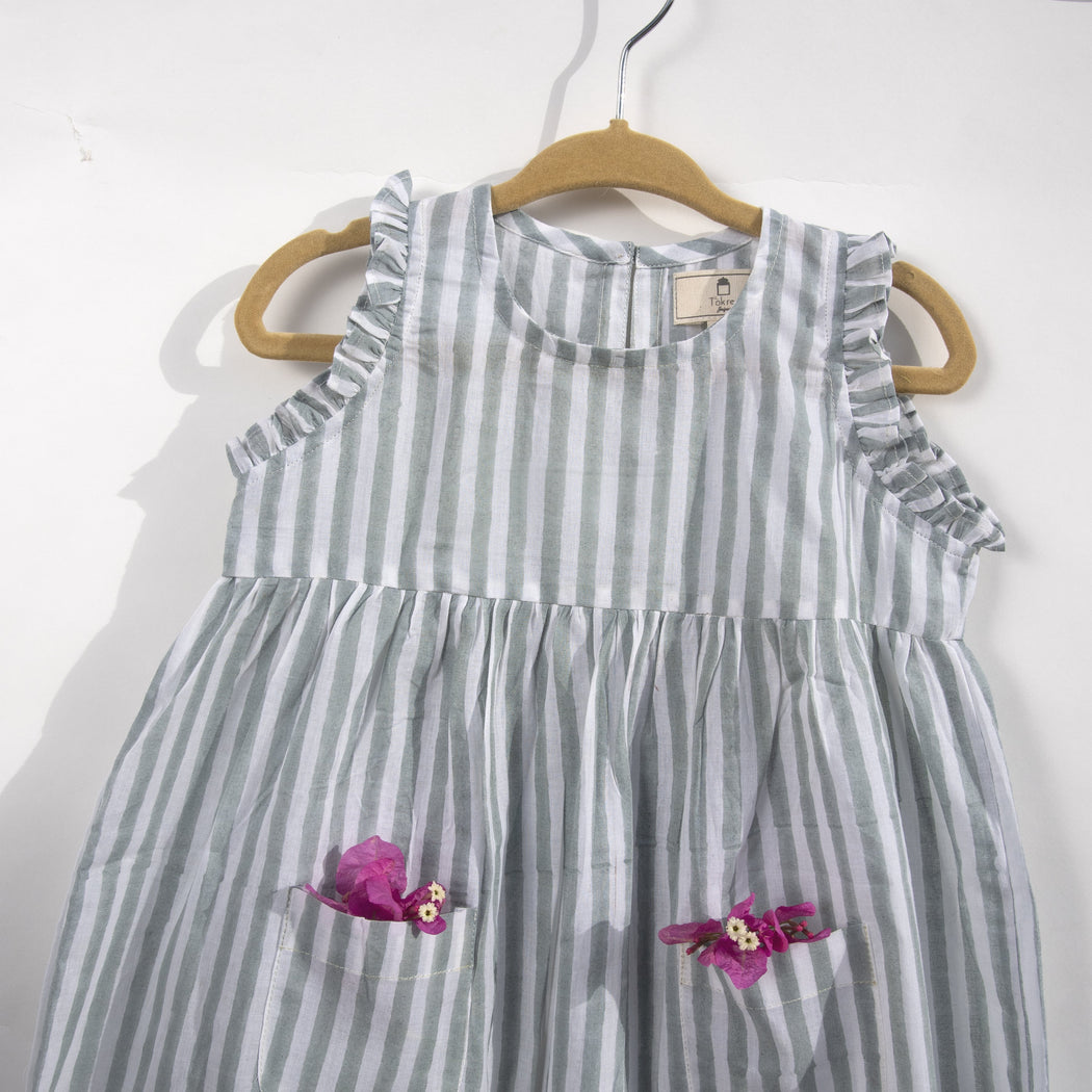 A white cotton mulmul dress for kids with hand-block printed pigeon grey stripes. This mid length dress has front pockets and a back button fastenings accentuated with ruffles on the sleeve, neck and pockets.